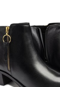 Botas de tobillo de cuero negro con textura suave, presentan acentos en forma de cremallera dorada y un tacón bajo en bloque. Vista en primer plano que resalta los detalles del diseño.