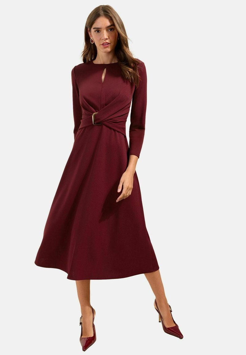 Kvinde iført en bordeaux rød midi-kjole med lange ærmer, nøglehulskæring, et snoet bælte og matchende spidse hæle.