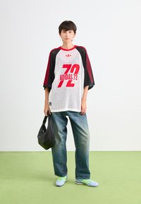 Camiseta oversized de malla blanca con gráfico rojo "ADIDAS 72", mangas negras, combinada con jeans azules holgados y un bolso negro. Zapatillas verdes brillantes.