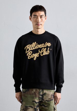 Mann in schwarzem Sweatshirt mit dem Schriftzug "Billionaire Boys Club" und Tarncargo-Hose, der vor einer schlichten hellgrauen Wand steht.