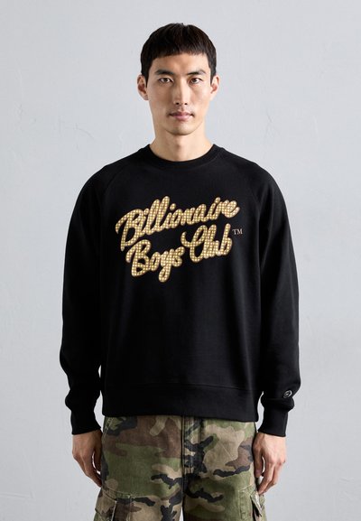 Man wearing black sweatshirt with "Billionaire Boys Club" text and camouflage cargo pants standing against plain light gray wall.