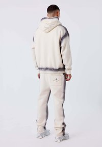 Uomo in piedi di spalle, che indossa un hoodie con sfumatura crema e grigia e pantaloni della tuta coordinati, con sneakers su uno sfondo neutro.
