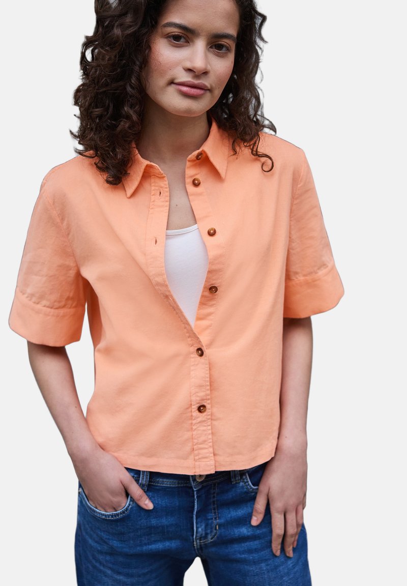 Jeune femme aux cheveux bouclés portant une chemise pêche à manches courtes déboutonnée par-dessus un haut blanc et un jean bleu, une main dans la poche.