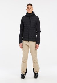 Schwarze isolierte Jacke mit Reißverschlusstaschen und Kapuze, kombiniert mit hellbeigen Hosen und schwarzen Skischuhen. Glatte, matte Textur.