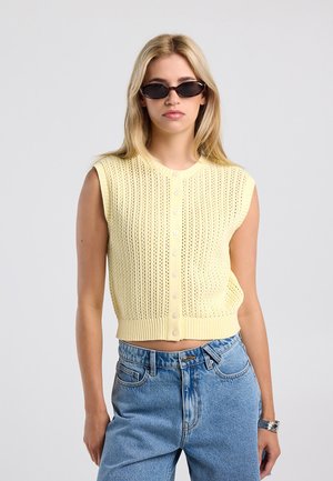 Vrouw met lang blond haar draagt een zwarte zonnebril, een mouwloos geel gebreid topje met knopen en een blauwe spijkerbroek, staand tegen een effen achtergrond.