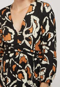 Robe noire avec un motif abstrait blanc et orange, décolleté en v, manches longues avec des poignets élastiques et un détail de jupe plissée.