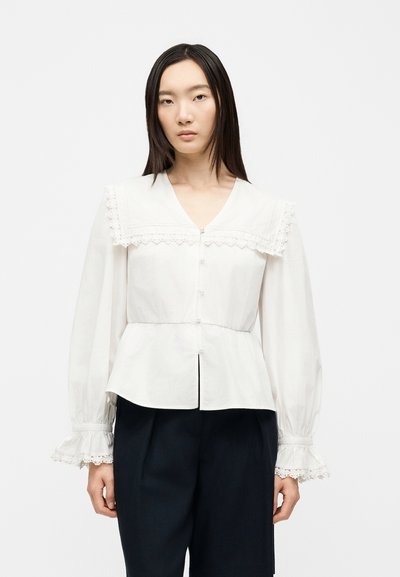 Femme portant un chemisier blanc avec bordure en dentelle au col et aux poignets, boutonné devant, et un pantalon noir, debout devant un fond uni.