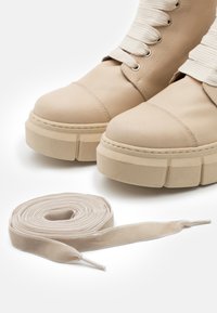 Botas de tornozelo em couro bege com uma sola grossa, apresentando um cadarço de tecido texturizado. Um cadarço de veludo enrolado na mesma cor encontra-se ao lado.