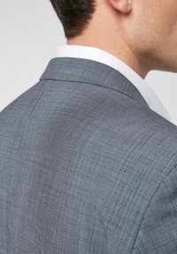 Grijze geruite blazer met een gestructureerde kraag, textuurstof en subtiel ruitpatroon; voorzien van een enkele split aan de achterkant en een gladde witte blouse eronder.