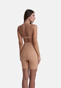 Donna con lunghi capelli scuri che indossa pantaloni shapewear a vita alta beige e un reggiseno abbinato, mostrata di spalle su uno sfondo bianco semplice.