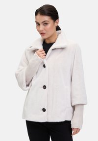 Jasno beżowa polarowa kurtka z dużymi czarnymi guzikami, szerokim kołnierzem i teksturowanymi mankietami; charakteryzuje się luźnym krojem i dwoma kieszeniami.