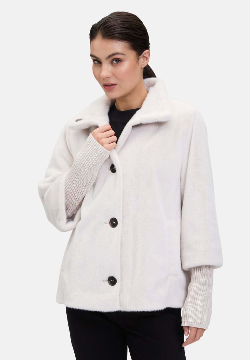 Jasno beżowa polarowa kurtka z dużymi czarnymi guzikami, szerokim kołnierzem i teksturowanymi mankietami; charakteryzuje się luźnym krojem i dwoma kieszeniami.