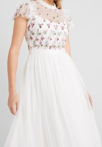 Valkoinen mekko, jossa on kukkakirjailtu bodysosetti paljetein, tüllihihoja ja teksturoidulla kankaalla varustettu hame, jossa on laskokset ja pilkkukuosi.