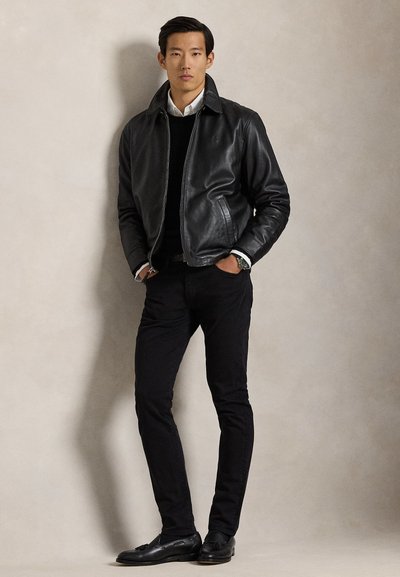 Homme debout contre un mur beige portant une veste en cuir noire, une chemise blanche, un pantalon noir et des mocassins noirs, les mains dans les poches.