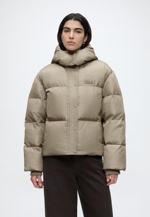 Femme portant une veste matelassée beige avec un col montant et des boutons-pression, associée à un pantalon foncé, debout devant un fond uni.
