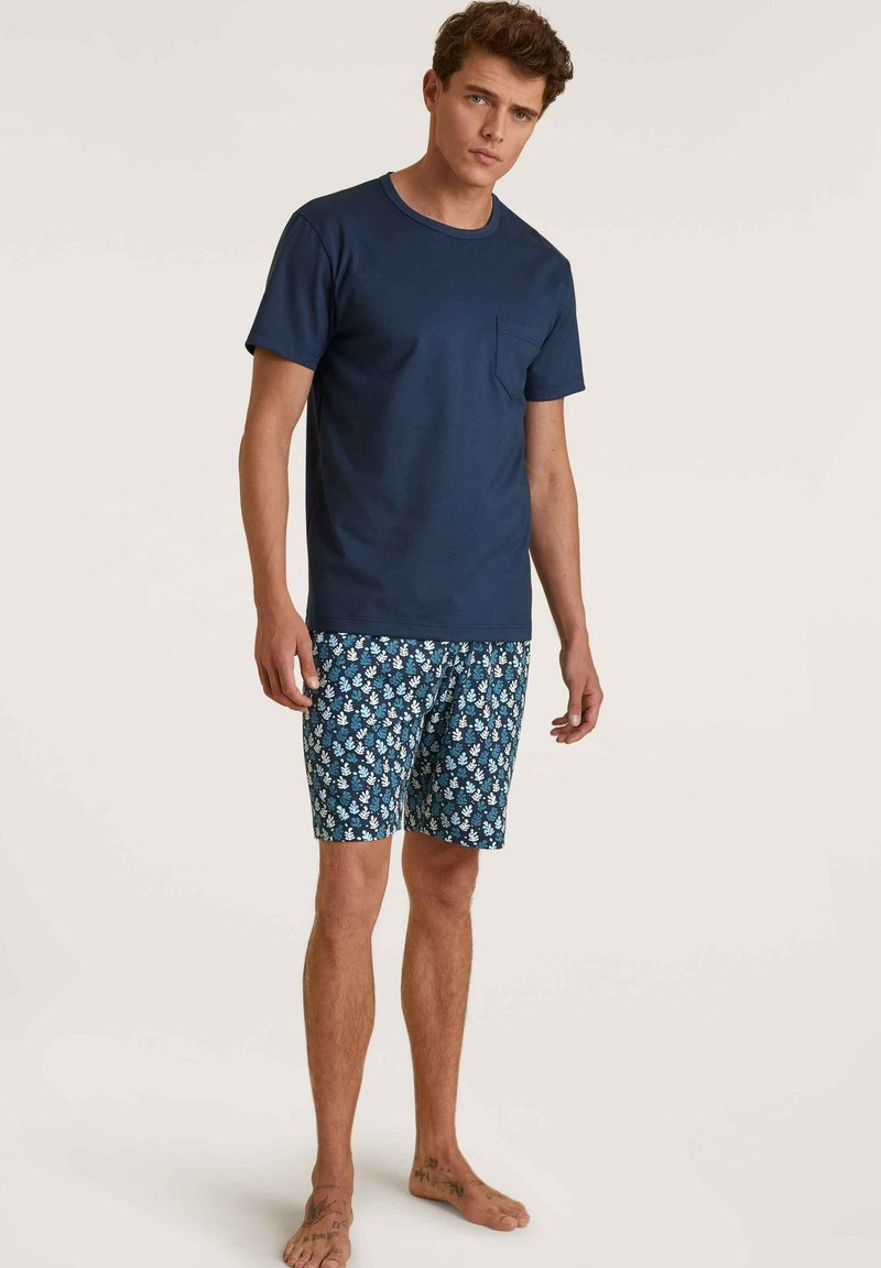 Marineblaues Baumwoll-T-Shirt mit einer Brusttasche, kombiniert mit gemusterten blauen Shorts mit weißen Blumenmustern. Das Modell ist barfuß.
