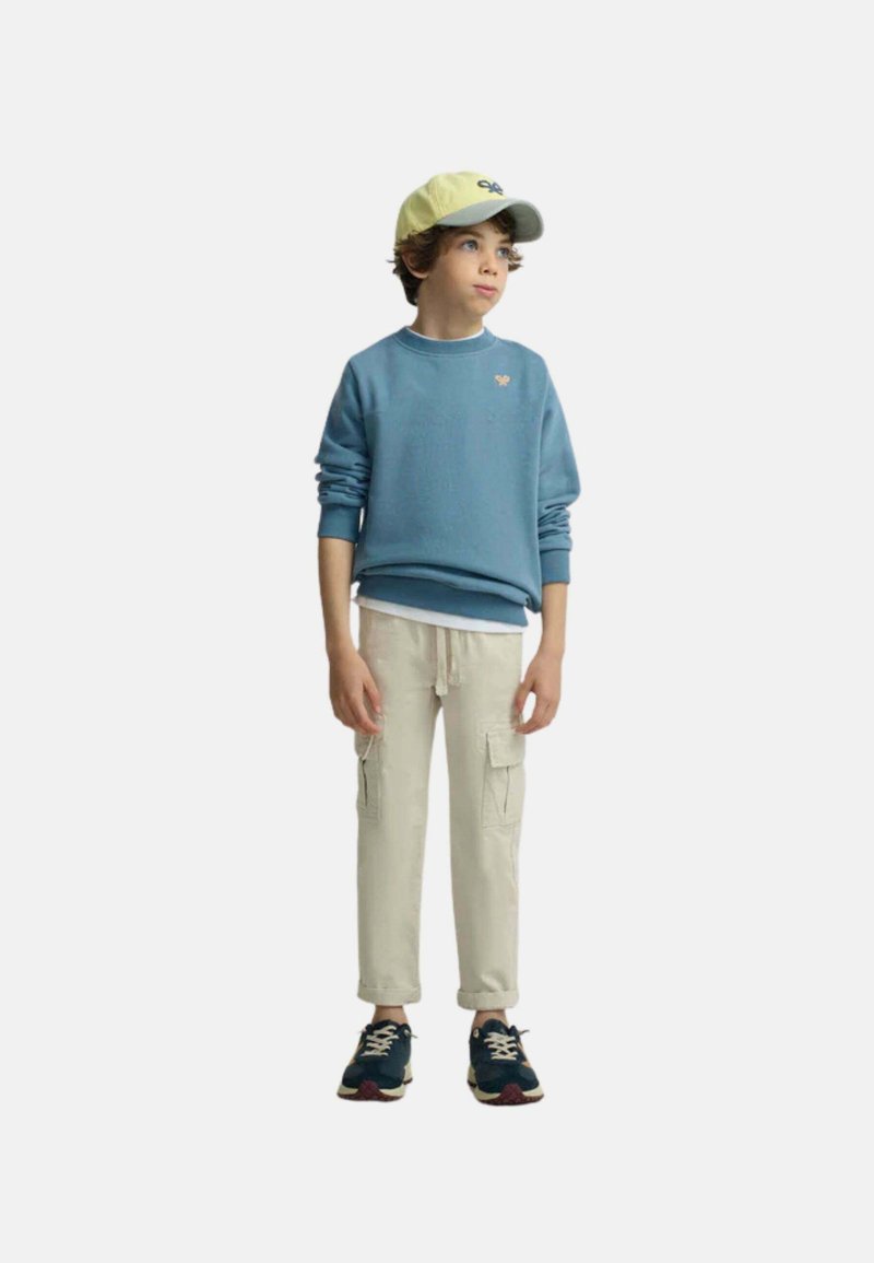 Niño con sudadera azul, pantalones cargo beige, deportivas azul marino y una gorra de béisbol amarilla y gris, de pie y mirando hacia un lado.