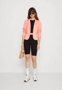 Korallinvärinen bleiseri, musta crop-top ja mustat pyöräilyshortsit. Pitää kädessään pientä beigeä laukkua. Valkoiset sukat ja lenkkarit. Tyylikkäät mustat aurinkolasit.