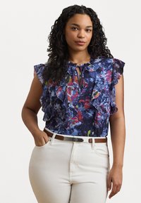 Bluse med blomsterprint som har volangermed, en knytehals og en marineblå bakgrunn med fargeklatter, kombinert med hvite høytlivsbukser.