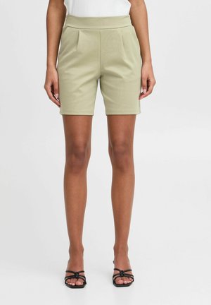 Tailored, knee-length shorts in light green fabric with two front pleats; paired with black strappy sandals. Smooth texture, fitted design.