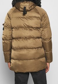 Chaqueta de plumas marrón con diseño acolchado horizontal, capucha forrada de pelo y una cinta de acento negra en la parte trasera. Textura suave y brillante.