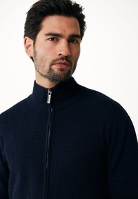 Námořnický svetr na zip s texturovaným vzorem, vysokým límcem a stříbrným zipem. Materiál vypadá jako pletenina a je přiléhavý.