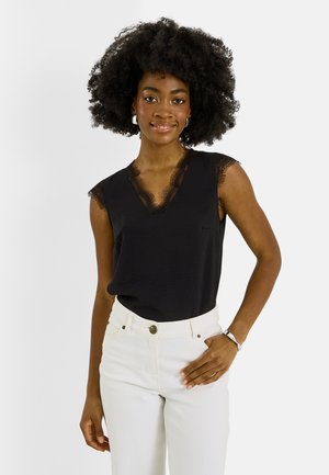 Femme aux cheveux bouclés portant un haut noir sans manches bordé de dentelle et un pantalon blanc, souriante avec une main sur la hanche.