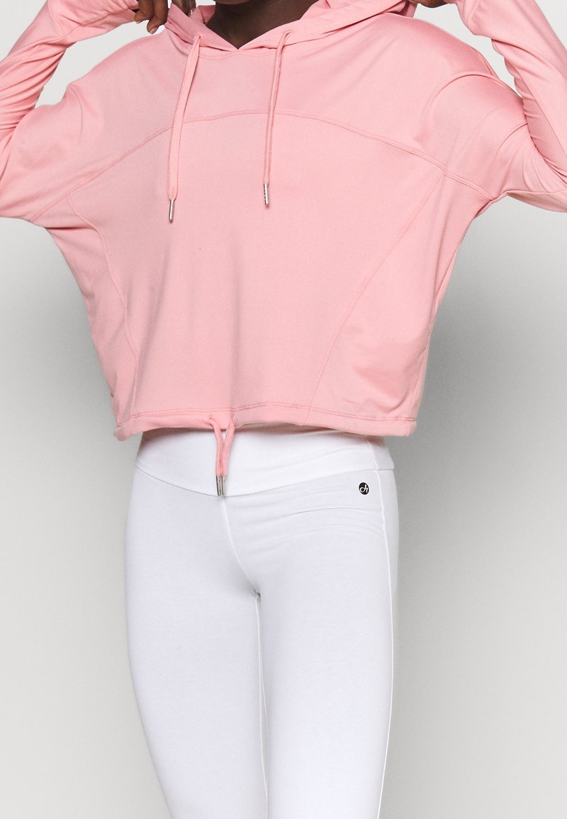 Hoodie court rose avec capuche, cordon de serrage et détails en métal argenté. Associé à des leggings blancs ajustés, mettant en valeur la texture et la coupe.