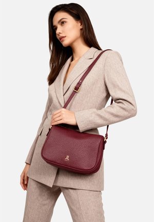 Borsa a tracolla in pelle color bordeaux con trama a grana, dettagli in metallo dorato e tracolla regolabile, tenuta da una persona con un outfit beige su misura.