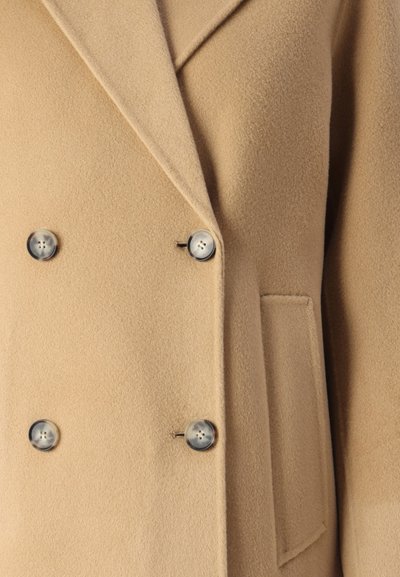 Cappotto doppiopetto color tan realizzato in tessuto morbido, con grandi bottoni e colletto a rever. Include una tasca laterale per un uso pratico.