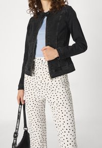 Žena oblečená v černé džínsové bundě přes světle modrý top, bílé kalhoty s černými puntíky a držící popruh černé kabelky.