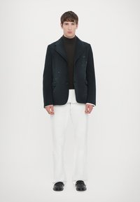 Blazer negro con salpicaduras de pintura visibles sobre un suéter de cuello alto marrón, combinado con pantalones anchos blancos y zapatos negros. Diseño simple y moderno.