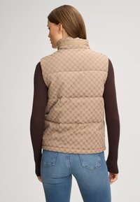 Beige gesteppte Weste mit Logo-Muster, hohem Kragen und vorderer Polsterung. Getragen über einem braunen Langarmoberteil und blauen Jeans.