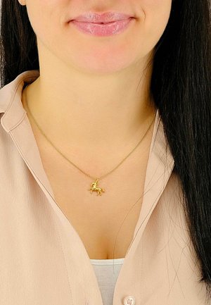 Pendentif licorne en or sur une chaîne fine, présentant des détails complexes. Le collier est porté sur un chemisier beige clair à col.