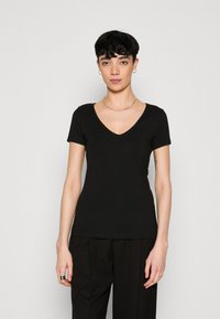 T-shirt en coton noir avec un décolleté en V profond et des manches courtes. Texture lisse et design ajusté, porté avec un pantalon noir ample.