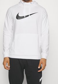 Homme portant un sweat à capuche blanc Nike avec logo swoosh noir et un pantalon noir avec un swoosh Nike blanc sur la jambe.