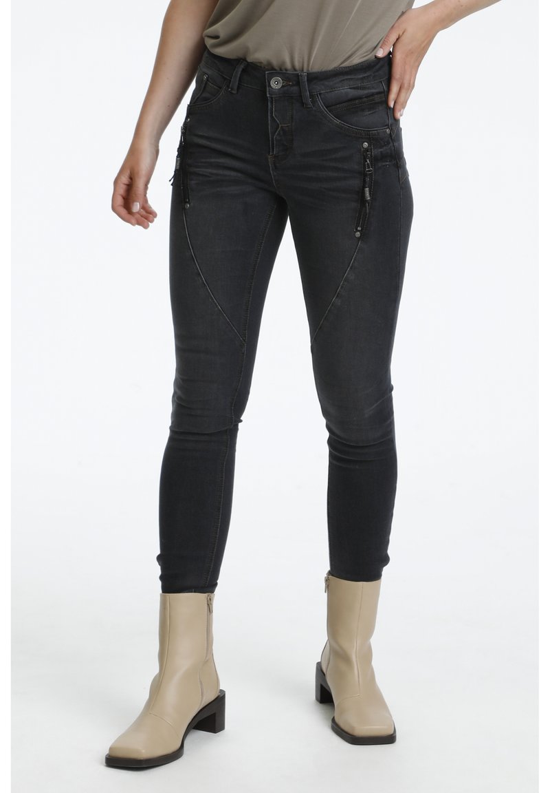 Jean skinny noir taille haute avec poches zippées, coutures discrètes et texture denim lisse, associés à des bottines beige à la cheville.