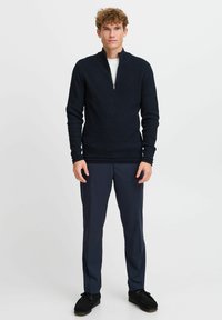Marineblauer Zip-Kragen-Pullover aus strukturiertem Stoff, mit gerippten Bündchen und Saum, kombiniert mit dunklen Hosen und schwarzem Schuhwerk. Einfaches, klares Design.
