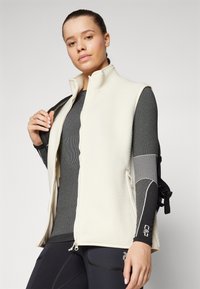 Fleece-Weste in Creme mit hohem Kragen und Reißverschluss, über einem grauen Langarmshirt getragen. Schwarze Leggings mit sichtbaren Akzentdetails.