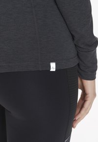 Pitkähihainen, tummanharmaa urheilu paita, jossa on kohokuvioitu kangas ja helmarekki, yhdistettynä mustiin legginsiin, joissa on kuviollinen sivupaneeli.