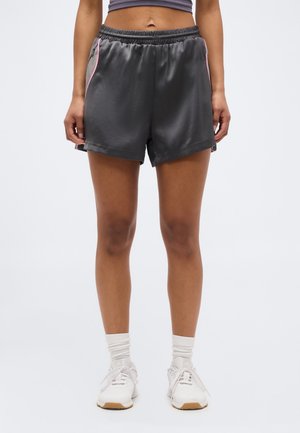 Žena oblečená v sivých športových šortkách s ružovým lemovaním, biele ponožky a biele bežecké tenisky stojaca na jednofarebnom pozadí.