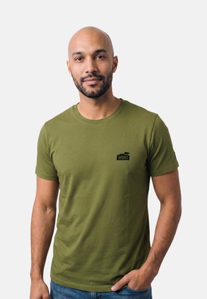 Uomo con barba corta e testa rasata indossa una maglietta verde oliva con piccolo logo nero "works" e jeans blu, in piedi rilassato.