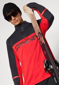 Rode en zwarte langetermijn-ski-top met een halve rits, met een logo op de borst, gecombineerd met een ski en skibindingen. Zwarte beanie en zonnebril.