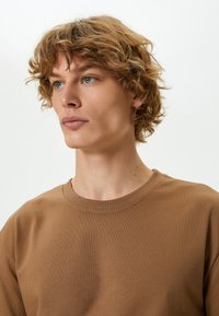 Brun sweatshirt crewneck med ribbet hals, laget av et mykt stoff. Sett på en modell med teksturert, bølgete hår og nøytral bakgrunn.