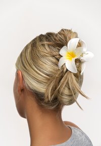 Vit blomma hårspänne i en blond knut, med mjuka kronblad och ett gult centrum, som visar en naturlig, elegant design mot en enfärgad bakgrund.