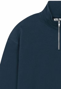 Sweat-shirt bleu marine avec col montant et une fermeture éclair argentée d'un quart de longueur. Texture douce avec des manches style raglan. Étiquette de la marque visible.