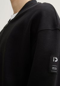 Giacca nera con zip, in tessuto testurizzato, colletto a costine e etichetta con marchio sulla manica. La vista ravvicinata mette in evidenza le cuciture e i dettagli delle spalle.