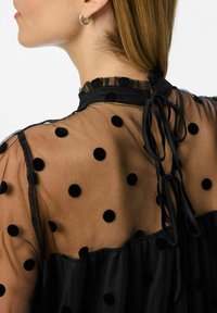 Crna bluzica s točkicama od prozirnog materijala, koja sadrži nabran ovratnik, vezanje vrpcom i mekanu crnu donju sloj.