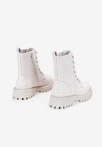 Stivali alla caviglia in pelle bianca con suola spessa, chiusura con lacci frontale e dettaglio con zip laterale. Texture liscia con punta rotonda e targhetta per una facile calzata.