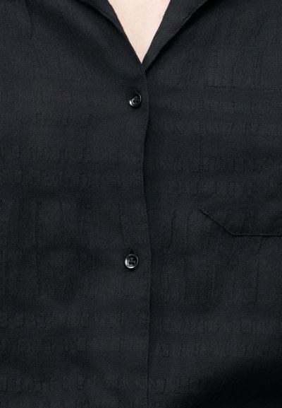 Nærbilde av svart skjorte med knapper, subtilt teksturert mønster og en lomme på høyre side av brystet.
